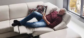 Mann entspannt auf hellem Stoffsofa mit elektrischer Relaxfunktion, gemütliches Wohnzimmer mit großen Fenstern und floralem Kissen Design.